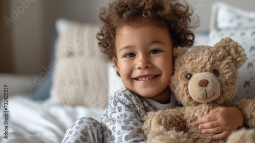 A young child with curly hair wearing pajamas smiling and holding a large teddy bear sitting on a bed with patterned pillows.