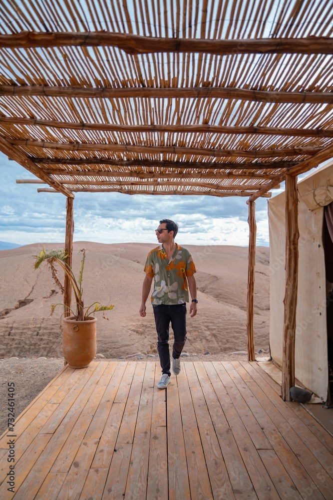 Fototapeta premium Man with sunglasses walks on a wooden deck in a desert setting. Agafay desert, Morocco.