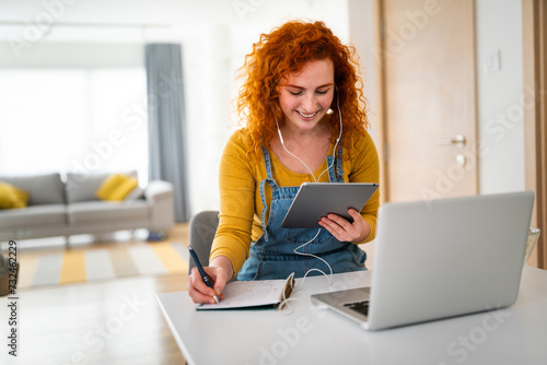 Relaxed, enthusiastic young college girl multitasking. Smiling woman writing in her notebook while looking at tablet and listening to music.