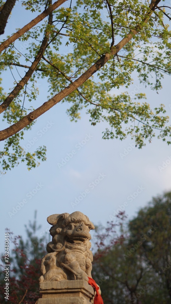 Fototapeta premium Weathered Asian stone statue resting atop a pedestal in a green, wooded area with trees
