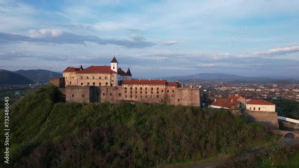 Mukachevo Castle on a high green hill in a warm evening sunset light with a beautiful blue sky aerial footage Mukachevo Castle on a high green hill in a warm evening sunset light with a beautiful blue sky aerial footage