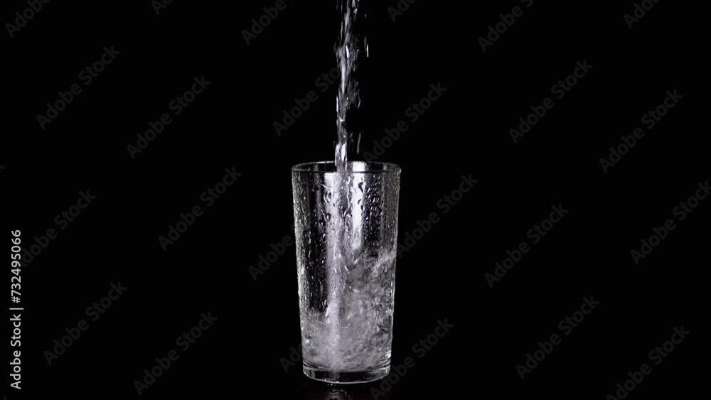 Pouring Pure Drinking Water into a Wet Glass of Water on a Black Background. Isolated. Slow motion. Stream of cold refreshing water pours into transparent glass with splashes. Water splash drops. Pouring Pure Drinking Water into a Wet Glass of Water on a Black Background. Isolated. Slow motion. Stream of cold refreshing water pours into transparent glass with splashes. Water splash drops.