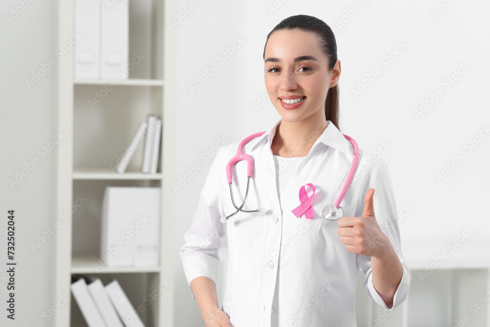 Mammologist with pink ribbon showing thumbs up in hospital, space for text. Breast cancer awareness