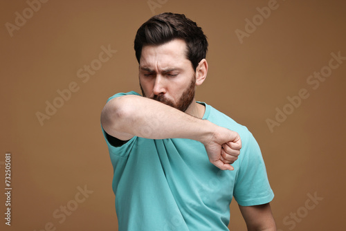 Sick man coughing into his elbow on brown background. Cold symptoms