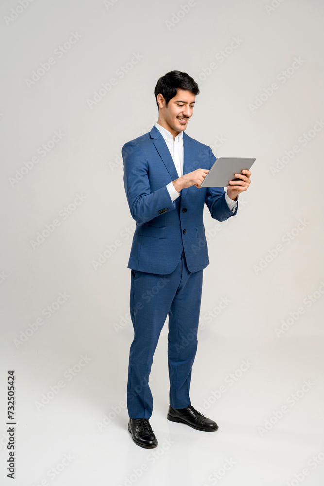 Young professional man in blue suit and white shirt is fully absorbed in reading a tablet screen while standing isolated on white. Concept of business, technology, and mobile connectivity. Full length Young professional man in blue suit and white shirt is fully absorbed in reading a tablet screen while standing isolated on white. Concept of business, technology, and mobile connectivity. Full length