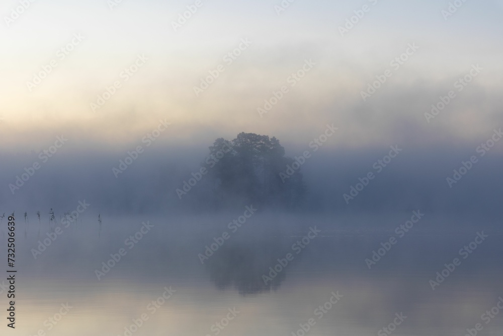 Fototapeta premium Tranquil lake surrounded by a lush green landscape with fog rolling in from the horizon.