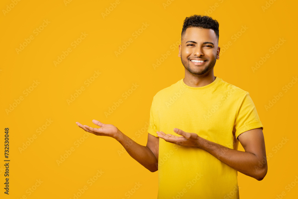 Look at here. Indian guy wearing casual t-shirt is advertising new product, points palms of both hands away at empty copy space, arab guy presents a novelty, isolated on yellow Look at here. Indian guy wearing casual t-shirt is advertising new product, points palms of both hands away at empty copy space, arab guy presents a novelty, isolated on yellow