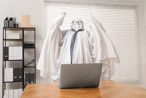 Overworked ghost. Man covered with white sheet using laptop at wooden table in office © New Africa