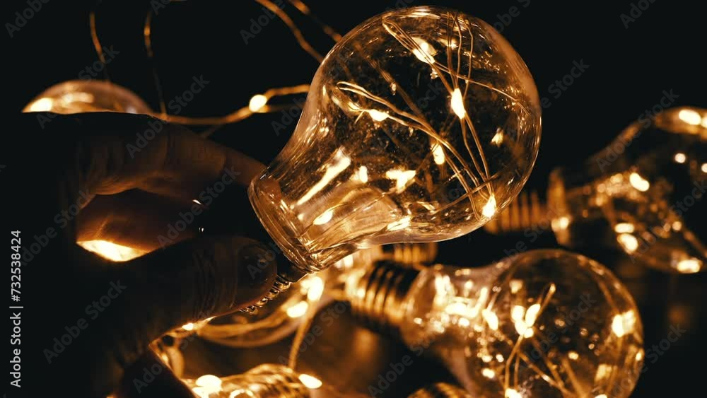 Hand Holds a Flickering Light Bulb on the Background of Glowing Light Bulbs. Warm light. Texture. Christmas decorations, vintage electric light bulbs in retro style illuminate the dark space. Dark. Hand Holds a Flickering Light Bulb on the Background of Glowing Light Bulbs. Warm light. Texture. Christmas decorations, vintage electric light bulbs in retro style illuminate the dark space. Dark.