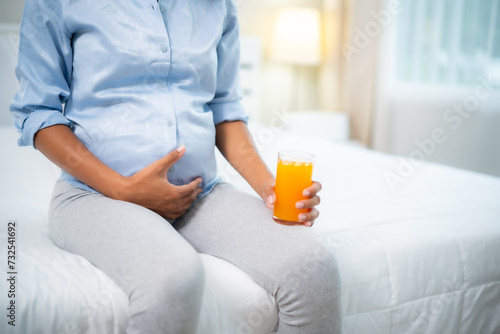 Crop of Young African American pregnant woman holding a glass of orange juce with smile of happiness while hand touching belly at home, taking care, healthy food , healthy concept with copy space. photo