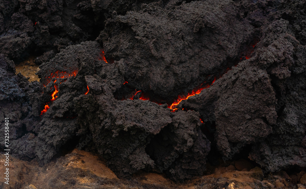 The quickly cooled lava crust creates an abstract. orange bright molten lava flowing on gray lava field and glossy rocky ground near volcano with fog in background. The quickly cooled lava crust creates an abstract. orange bright molten lava flowing on gray lava field and glossy rocky ground near volcano with fog in background.