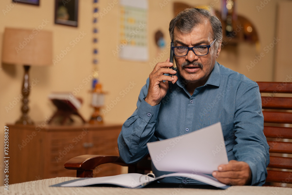 Indian senior man talking or speaking on mobile phone by holding documents at home - concept of consultation, financial management and consultation