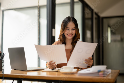 A young professional woman smiles while examining papers in a well-lit, modern office setting, with a laptop. © Wasana