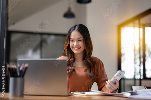 A focused businesswoman using a smartphone while working on a laptop at a desk, in office space.