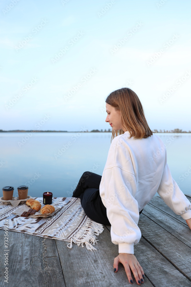 the girl sits on a wooden pier near the lake. aesthetic photo. picnic on a wooden pier with coffee and croissants