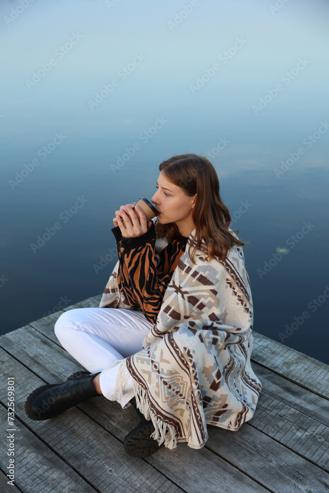 Fototapeta premium the girl is sitting on a wooden pier near the lake wrapped in a blanket and drinking coffee. aesthetic photo. autumn vibe