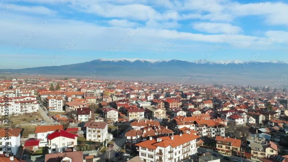Drone flies sideways over Bansko while facing beautiful Rila Mountains covered in snow, early morning shot in winter.