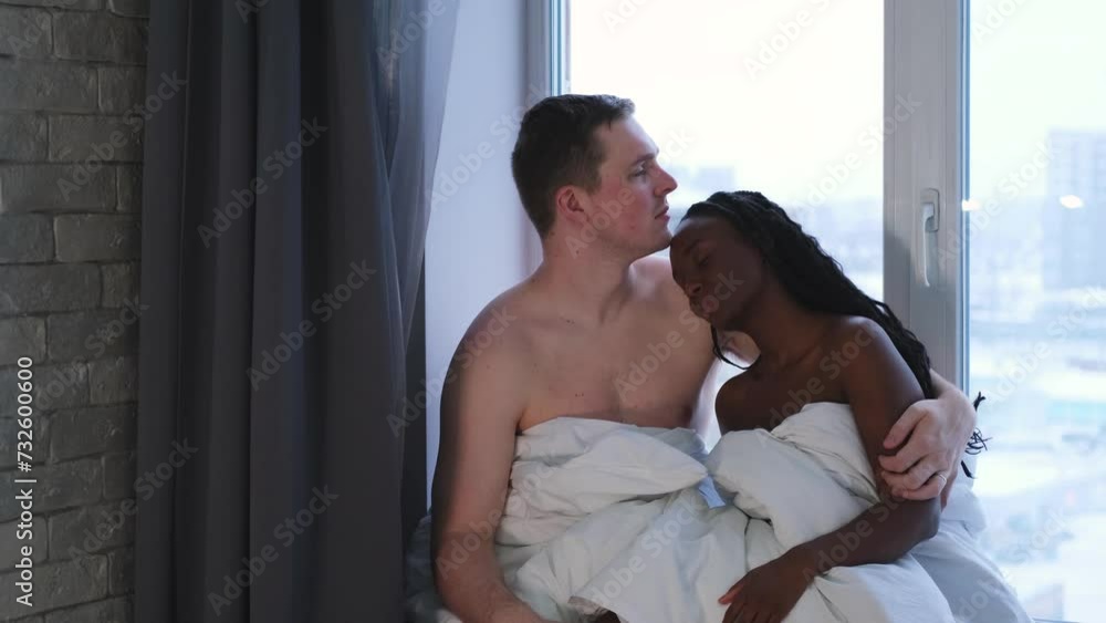 Happy loving couple wrapped in a white warm blanket hugging while sitting on the windowsill on a cloudy day. A beautiful wife and a handsome husband look at each other, enjoy a tender moment. Happy loving couple wrapped in a white warm blanket hugging while sitting on the windowsill on a cloudy day. A beautiful wife and a handsome husband look at each other, enjoy a tender moment.