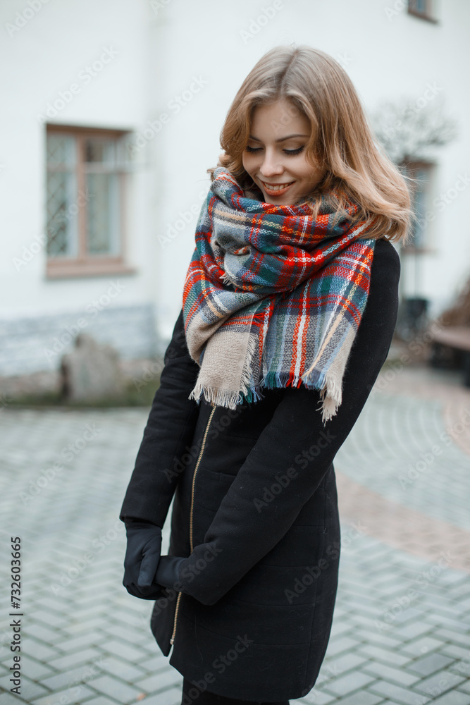 Beautiful happy woman with a sweet smile in a fashionable black coat with a checkered scarf walks in the city Beautiful happy woman with a sweet smile in a fashionable black coat with a checkered scarf walks in the city