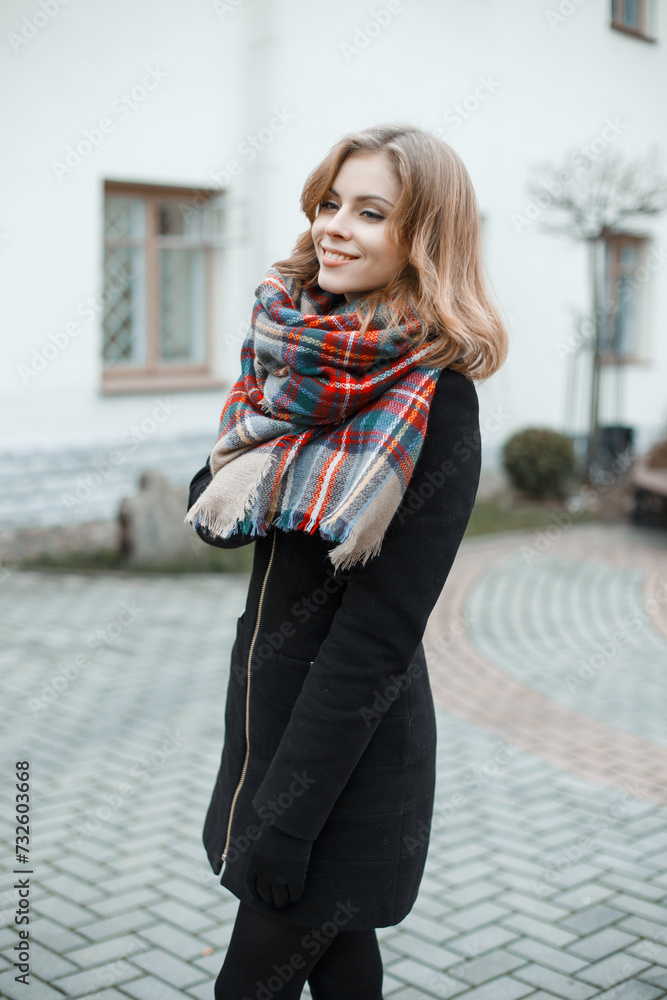 Stylish beautiful young woman model with a smile with a fashion vintage checkered scarf in a black coat walks on the street
