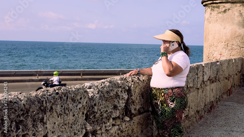 An elderly woman standing at the edge of the Cartagena city walls, dressed casually with a visor, looking out towards the sea and talking on the phone