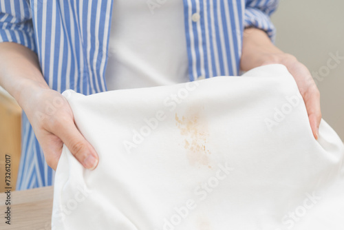 Housewife concept, frustrated young maid woman hand holding white t-shirt, showing make cloth stain, spot dirty or smudge, sauce spill, splash on clothes, dirt stains for cleaning before washing.