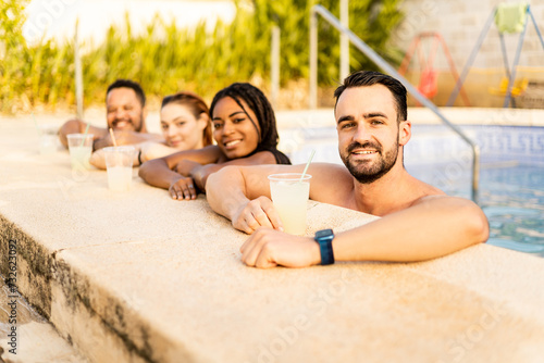 A group of 4 friends of different ethnicity and physique are in a swimming pool in a country house. The young people are looking at the camera happily leaning on the edge of the pool