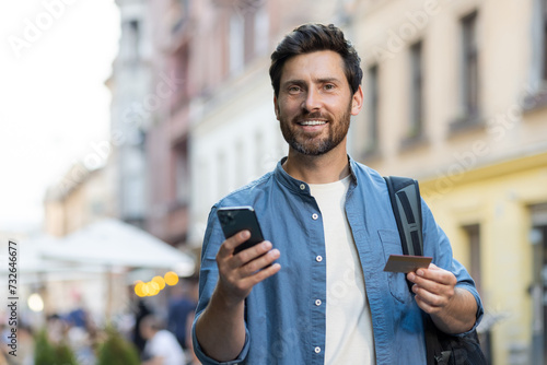 Portrait of a young male tourist standing on a city street with a backpack, holding a credit card and a mobile phone. Smiling and looking at the camera
