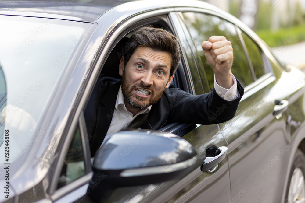 An angry and irritated young man in a business suit looks out of the car window. He is behind the wheel, shouting at the camera and waving his hands threateningly. Close-up photo
