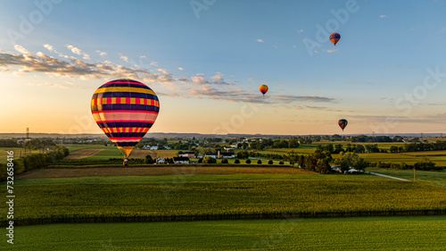 An Aerial View of a Vividly Striped Hot Air Balloon Floating Close To The Ground With Others In The Distance Above Lush Green Farmlands Under A Softly Lit Sky.