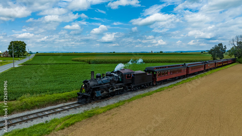 Vintage Steam Locomotive Pulling Red Passenger Cars Alongside Lush Fields And A Dirt Path Under A Broad Sky With Fluffy Clouds.