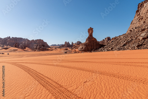 Fototapeta Naklejka Na Ścianę i Meble -  Landscape of the Red Tadrart in the Sahara Desert, Algeria. Tire tracks in the sand in front of a sandstone peak reminiscent of the football World Cup trophy