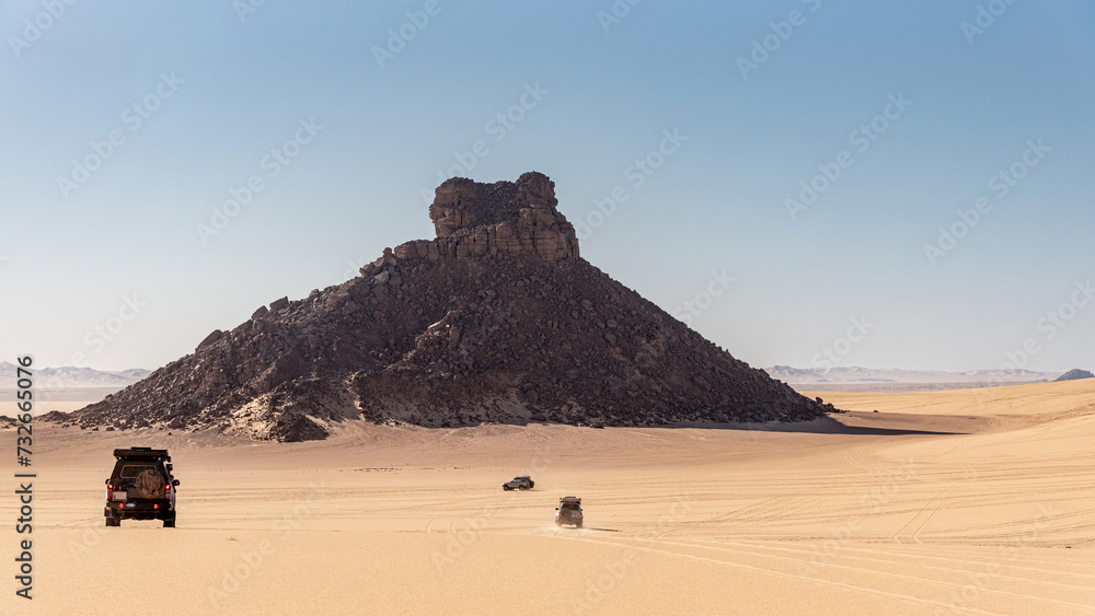 Landscape of the Tiboukaine region in the Sahara Desert, Algeria. The jeeps head towards a mountain of scree that seems to spring from the sand. Landscape of the Tiboukaine region in the Sahara Desert, Algeria. The jeeps head towards a mountain of scree that seems to spring from the sand.