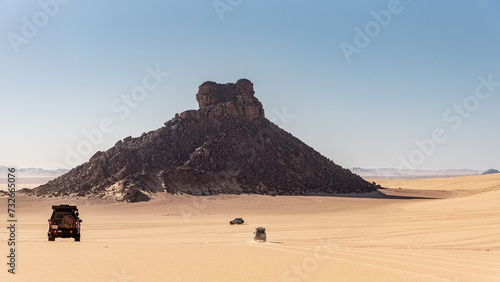 Landscape of the Tiboukaine region in the Sahara Desert, Algeria. The jeeps head towards a mountain of scree that seems to spring from the sand.