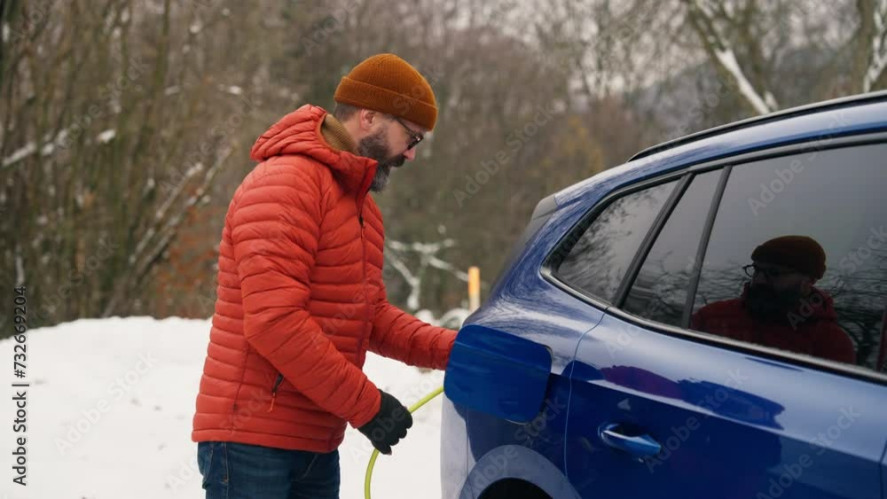 Man charging electric car during cold snowy day. Side view of hansome man putting charger in charging port during winter, cold weather. Charging and driving electric vehicles during winter season. Man charging electric car during cold snowy day. Side view of hansome man putting charger in charging port during winter, cold weather. Charging and driving electric vehicles during winter season.
