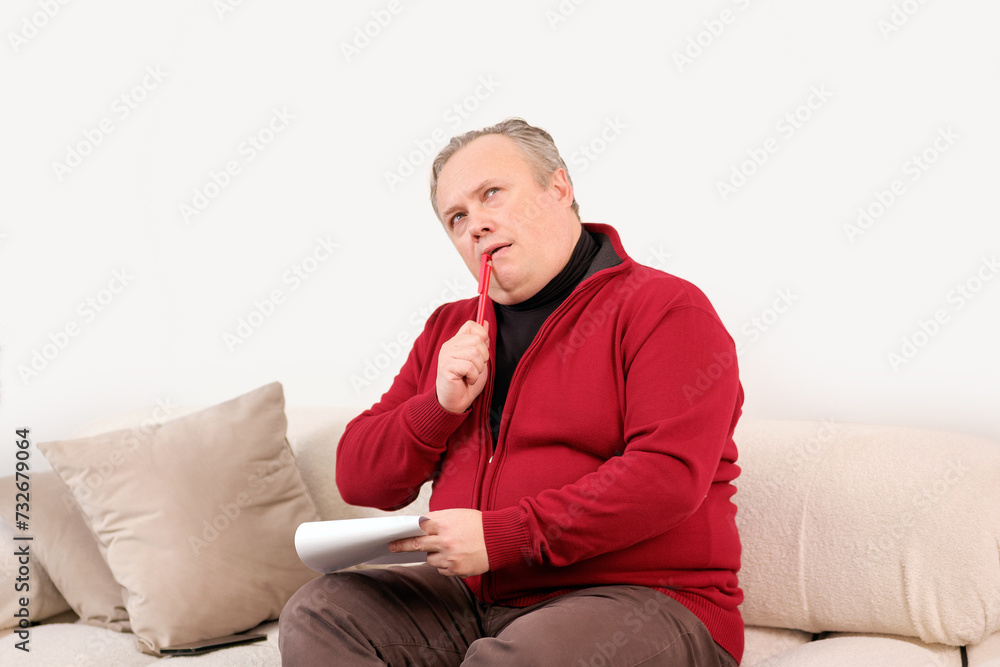 man thinks intently before writing in a paper notebook while sitting on the sofa man thinks intently before writing in a paper notebook while sitting on the sofa