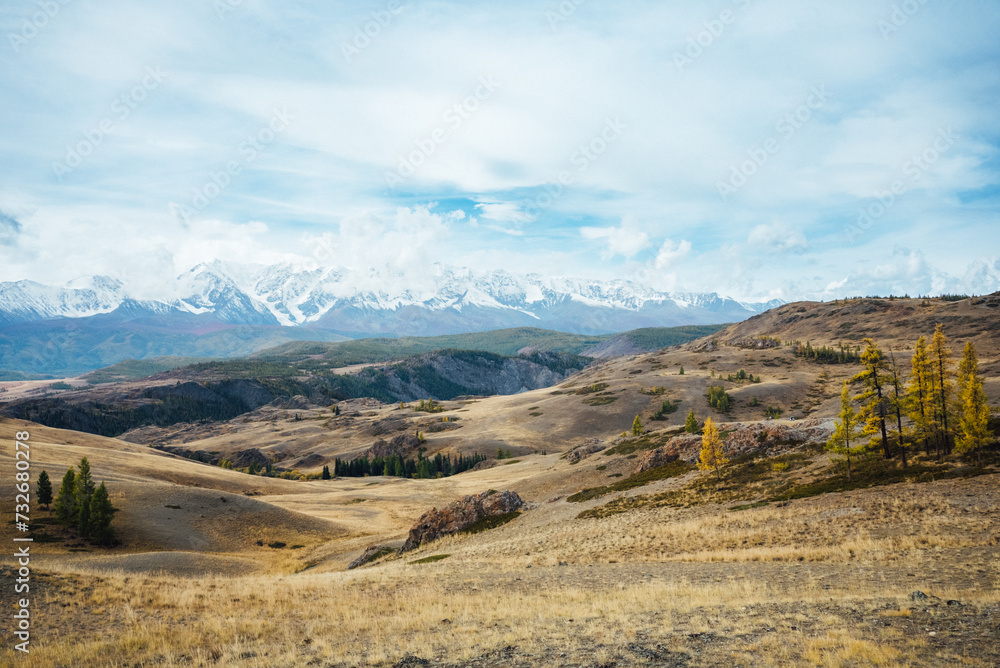 Expansive view of a vast valley with rolling hills and sparse vegetation, leading to a majestic mountain range capped with snow under a serene blue sky with scattered clouds. Stunning mountain