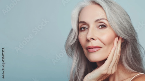 close-up portrait of an elegant senior woman with gray hair, gently touching her face and smiling subtly, set against a neutral background, suggesting a skincare or beauty theme.