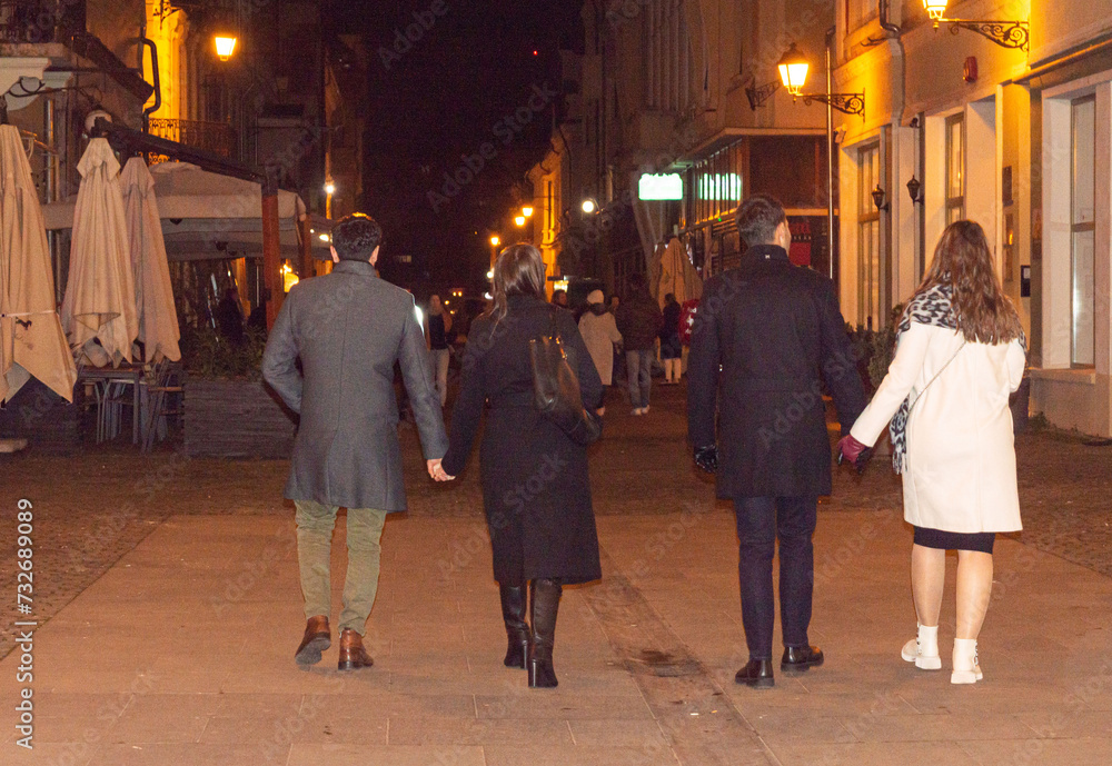 Two fashionable couples holding hands wearing elegant coats walking on the street at night in the city square. Rear view. Two couples meeting in the evening in the city. Urban landscape at nighttime
