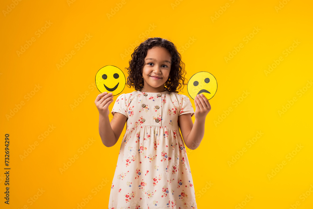 Smiling kid girl holding sad and happy emoticons in hands. Mental health, psychology and children's emotions concept - obrazy, fototapety, plakaty 