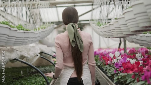 A young pretty girl with a kerchief on her hair walks between the rows of greenery and flowers in a greenhouse. The camera follows the girl behind. Video 4k, rec 709