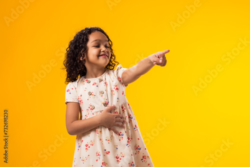 Kid girl mocking and teasing at someone showing finger at camera and holding stomach over yellow background. Bulling concept photo