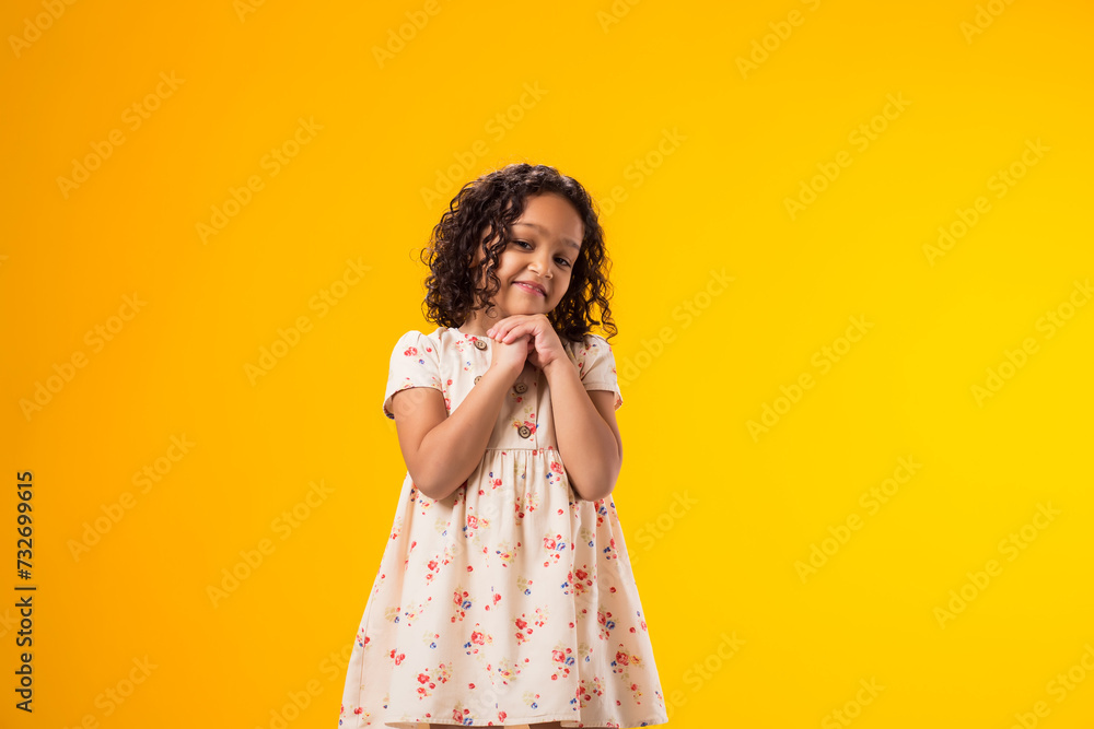 Smiling kid girl holding hands together under face looking at camera over yellow background. Childhood and positive emotions concept Smiling kid girl holding hands together under face looking at camera over yellow background. Childhood and positive emotions concept