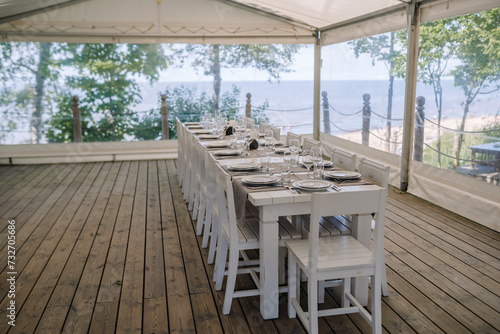 Jurmala, Latvia - july 25, 2023 - Outdoor dining setup under a tent on a wooden deck with a long table set for a meal, overlooking a beach and the sea. © Raivo