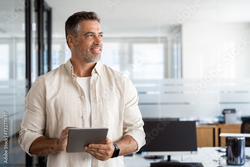 Happy Latin Hispanic bearded stylish mature adult professional business man, smiling Indian senior businessman CEO holding digital tablet using fintech tab application standing in office,looking aside