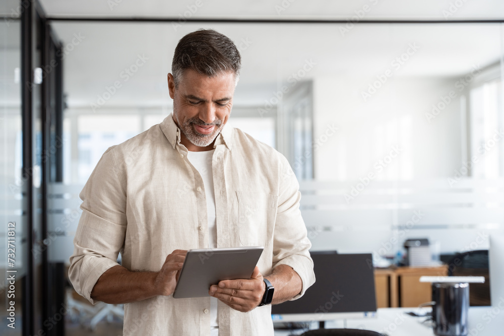 Happy Latin Hispanic bearded stylish mature adult professional business man, smiling Indian senior businessman CEO holding digital tablet using fintech tab application standing inside company office. Happy Latin Hispanic bearded stylish mature adult professional business man, smiling Indian senior businessman CEO holding digital tablet using fintech tab application standing inside company office.