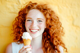 Portrait of a smiling curly redhaired woman by a yellow wall with eating a icecream cone