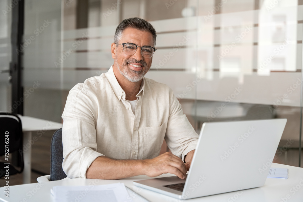 Portrait of mature Indian or Latin business man ceo trader using laptop computer, typing, working in modern office. Middle-age Hispanic smiling handsome businessman entrepreneur looking at camera. Portrait of mature Indian or Latin business man ceo trader using laptop computer, typing, working in modern office. Middle-age Hispanic smiling handsome businessman entrepreneur looking at camera.