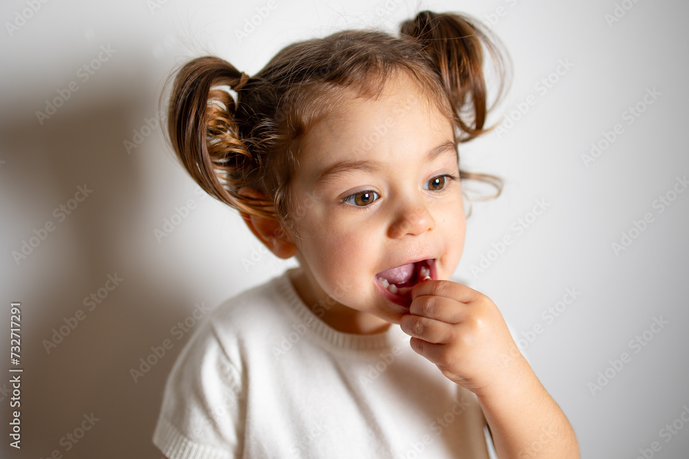 Close portrait cute caucasian brunette toddler girl with two tails in white dress smiling with teeth, Montessori method on white background 