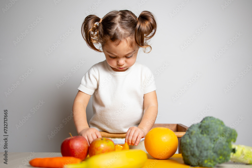 Cute caucasian brunette toddler girl with two tails in white dress staying at the learning tower and cutting apple, Montessori method on white background 